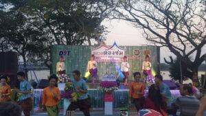 งานประเพณีไทยบนเวทีกลางแจ้งมีการแสดงนาฏศิลป์ดั้งเดิมไทย ผู้แสดงสวมใส่ชุดผ้าไหมสีทองและสีม่วงเข้มบนเวทีตกแต่งด้วยดอกไม้สีชมพูแดง
