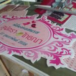 ป้ายอีเวนต์งานแต่งงานสีชมพูและขาวประดับลายอาร์ตโนวโวที่วางบนโต๊ะแสดงจัดเรียงพร้อมเอกสารและวัสดุโปรโมตต่างๆ