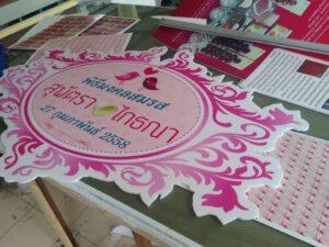 ป้ายอีเวนต์งานแต่งงานสีชมพูและขาวประดับลายอาร์ตโนวโวที่วางบนโต๊ะแสดงจัดเรียงพร้อมเอกสารและวัสดุโปรโมตต่างๆ
