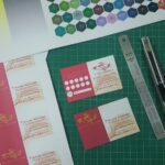 ชุดเอกสารการออกแบบที่มีบัตรธุรกิจสีชมพูและสีแดง ตัวอย่างสีหกเหลี่ยม และเครื่องมือออกแบบบนเสื่อตัดสีเขียว
