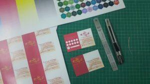 ชุดเอกสารการออกแบบที่มีบัตรธุรกิจสีชมพูและสีแดง ตัวอย่างสีหกเหลี่ยม และเครื่องมือออกแบบบนเสื่อตัดสีเขียว