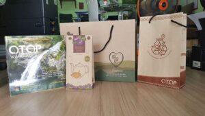 กลุ่มสินค้าและวัสดุการตลาดของ OTOP ที่วางบนโต๊ะไม้ รวมถึงนิตยสารสีเขียว ถุงกระดาษสีครีมพิมพ์ลายใจ และกล่องบรรจุภัณฑ์ OTOP