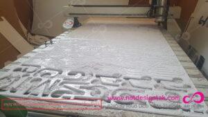 เครื่องตัดเลเซอร์ CNC กำลังแกะสลักอักษรและลวดลายทางศิลปะบนแผ่นวัสดุสีขาว ในสตูดิโอด้านข้างมีตัวอักษรแกะสลักจำนวนมากวางอยู่