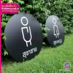 ป้ายทรงกลม 2 อันวางอยู่บนสนามหญ้า แทนห้องน้ำชายและหญิง พร้อมไอคอนเก๋ๆ และข้อความภาษาไทยน่าจะหมายถึง 'ป้ายสุขาชาย ห