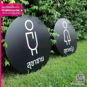 ป้ายทรงกลม 2 อันวางอยู่บนสนามหญ้า แทนห้องน้ำชายและหญิง พร้อมไอคอนเก๋ๆ และข้อความภาษาไทยน่าจะหมายถึง 'ป้ายสุขาชาย ห