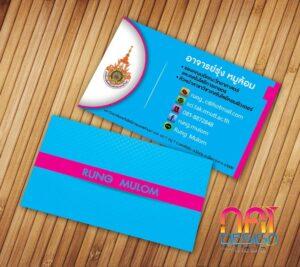 นามบัตรสีฟ้าและสีชมพูของรุ่งมูลม มีโลโก้วัดสีทองและข้อมูลติดต่อบนพื้นไม้